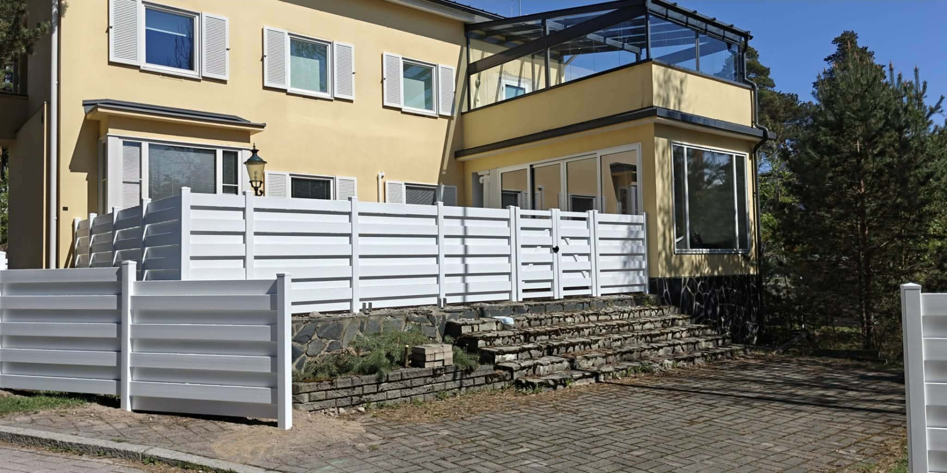 Valkoinen vinyyliaita ja portti keltaisen talon kiviterassin kohdalla.