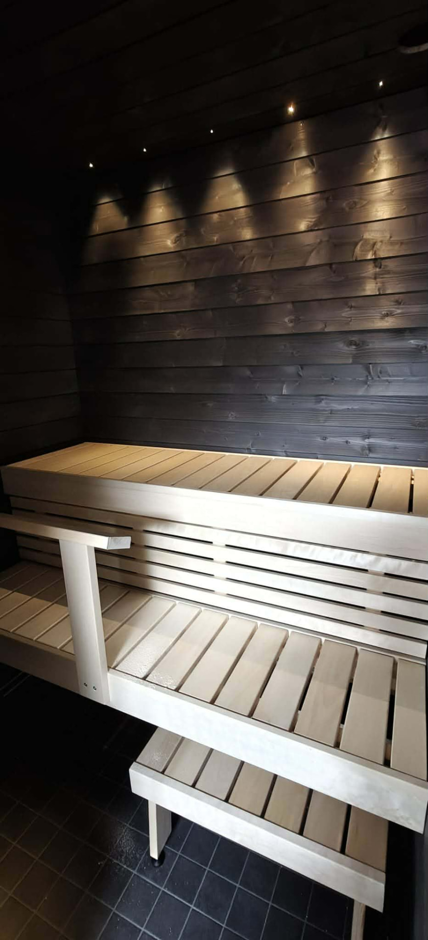 Tummasävyinen sauna, vaaleat lauteet ja kattovalaistus.